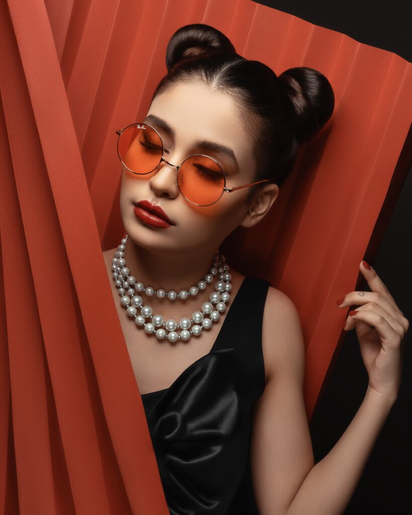 Asian girl wearing oversized sunglasses with orange tint lenses while wearing a little black dress and reminding of sunglasses history
